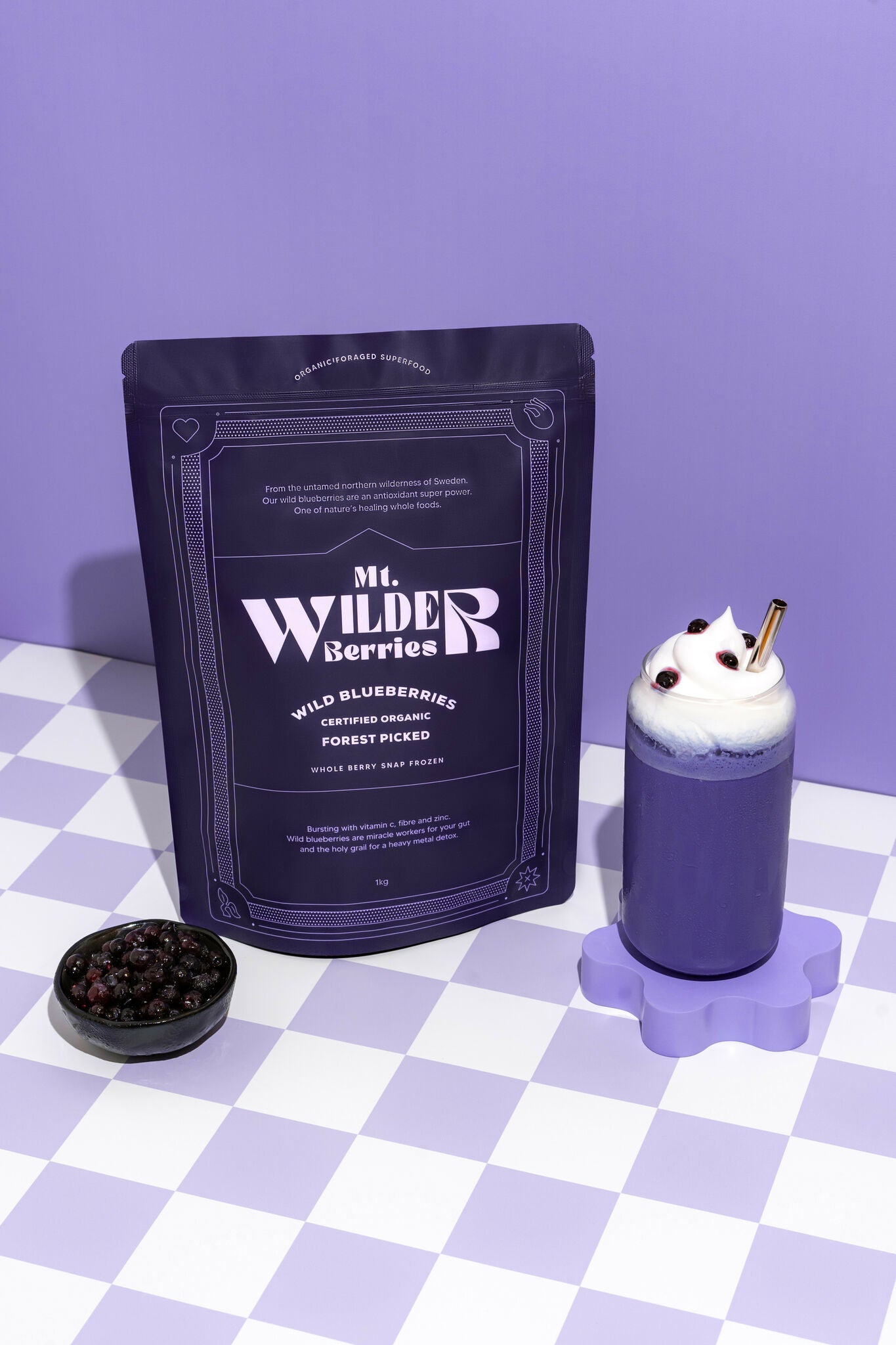 A package of 'Mr. Wilder Berries' certified organic wild blueberries, with a small bowl of blueberries in front and a prepared dish with blueberry sauce and whipped cream on a purple surface with a checkered pattern.
