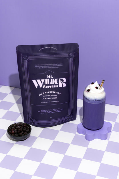 A package of 'Mr. Wilder Berries' certified organic wild blueberries, with a small bowl of blueberries in front and a prepared dish with blueberry sauce and whipped cream on a purple surface with a checkered pattern.