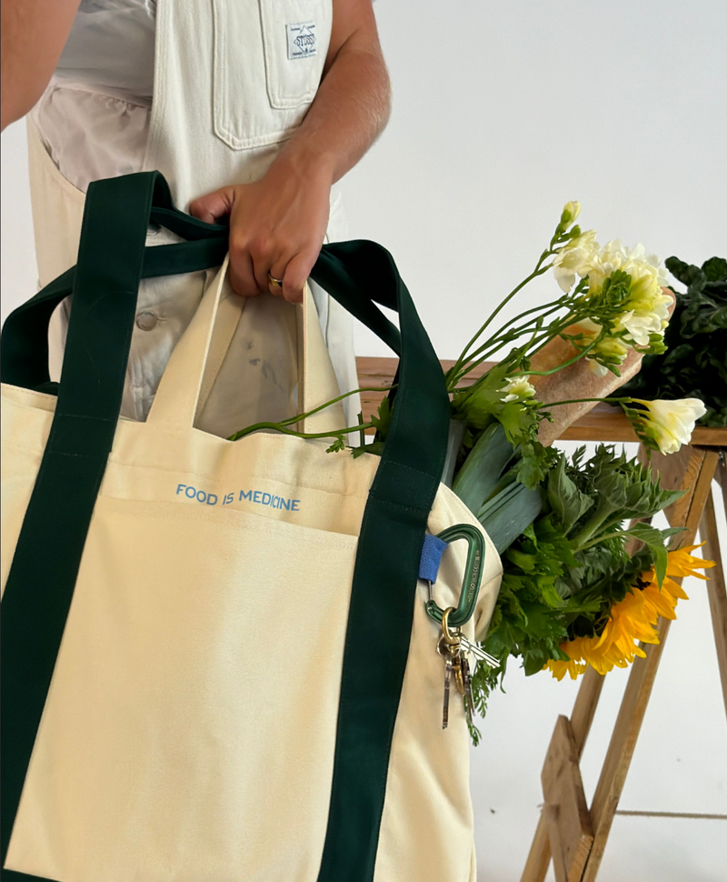 Person holding a beige tote bag with 'Food is Medicine' logo, filled with groceries and flowers.