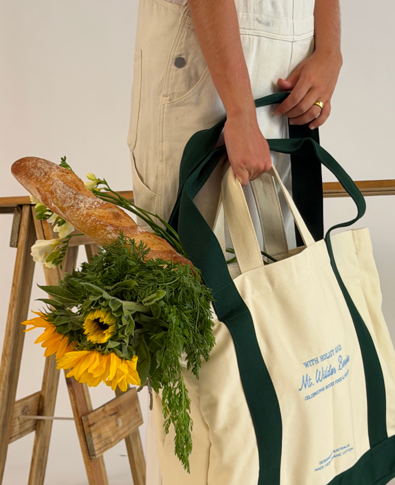 Person holding a tote bag with bread and flowers, standing next to a wooden ladder.