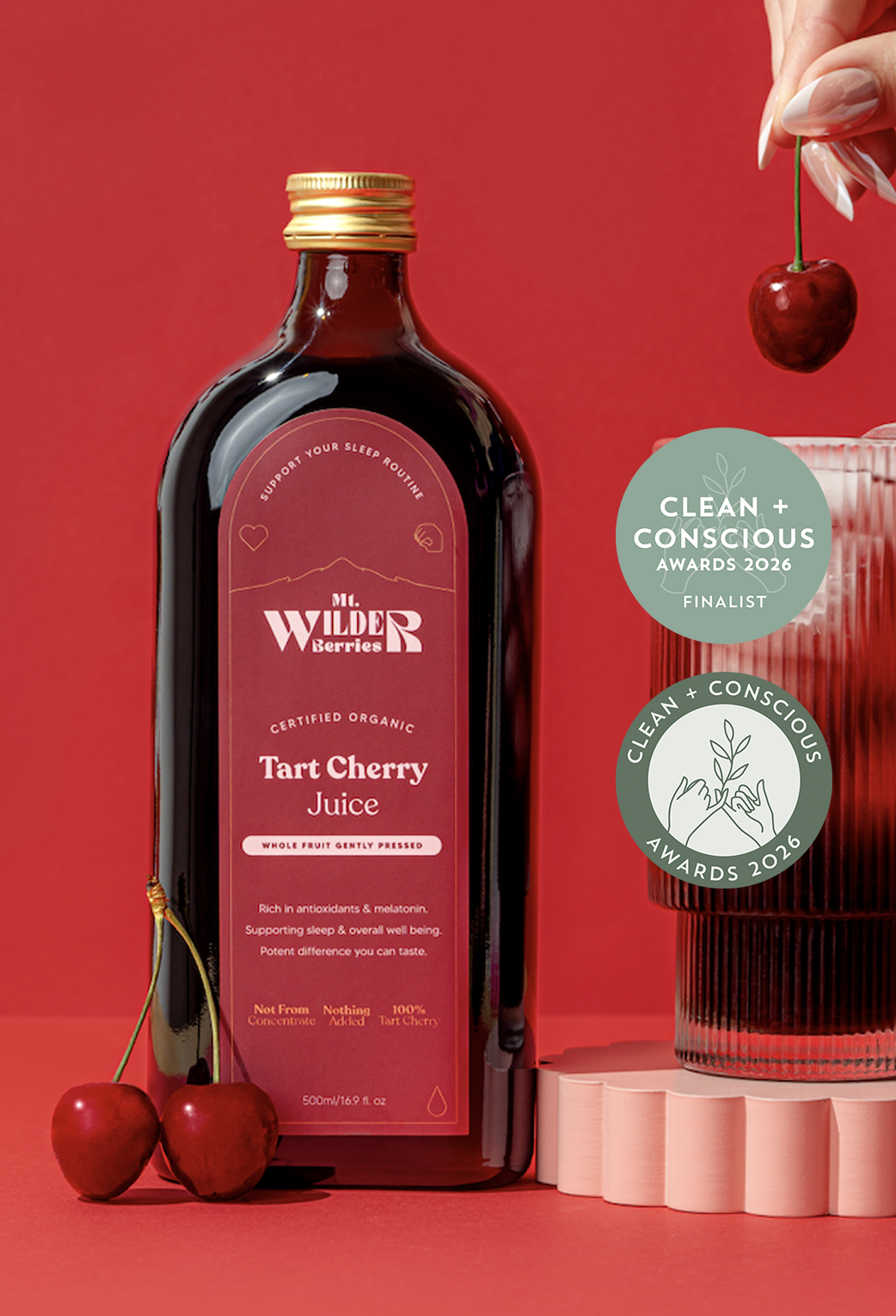 A bottle of certified organic tart cherry juice with a glass filled with ice and cherry juice, garnished with a cherry, on a red background, along with two cherries on the side.