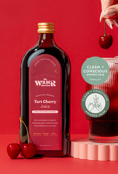 A bottle of certified organic tart cherry juice with a glass filled with ice and cherry juice, garnished with a cherry, on a red background, along with two cherries on the side.