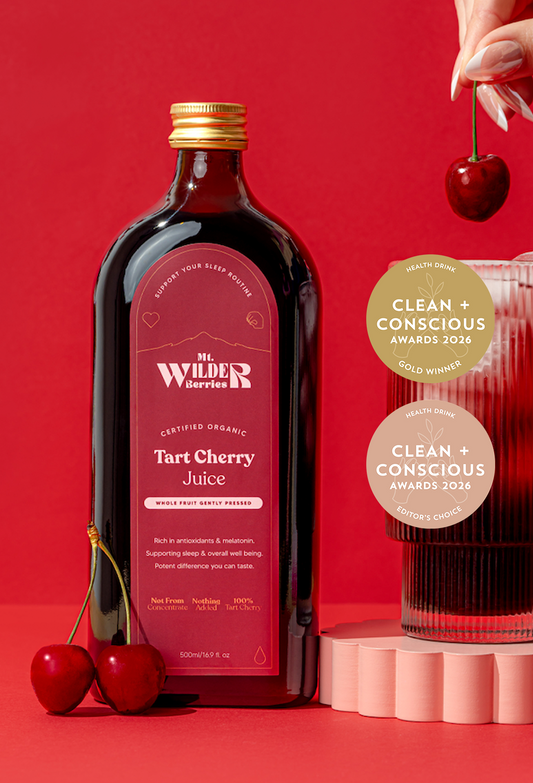 A bottle of Mr. Wilder Barriers Tart Cherry Juice stands on a red studio background with two whole cherries at the base and a hand holding a cherry at the top right.